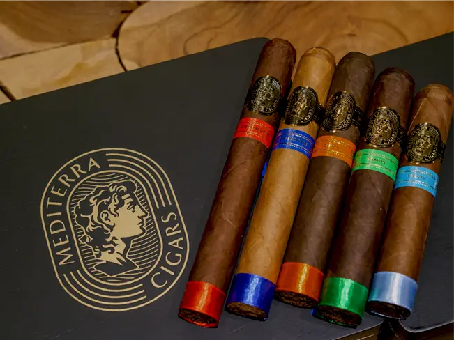 Five premium Mediterra cigars displayed on a dark branded presentation box featuring the gold Mediterra Cigars logo with a classical Greek-style profile. Each cigar showcases distinctively colored bands labeled Thessaloniki (red), Santorini (blue), Chios (orange), Samos (green), and Milos (sky-blue), highlighting the variety in wrapper shades and flavors.